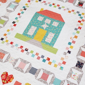 May include: A white quilt with a colorful house design in the center. The house is surrounded by a border of gray, white, and red squares. The quilt is decorated with hearts, flowers, and other geometric shapes.