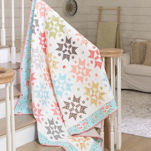 Pode incluir: Um edredom branco com um padrão de estrela em forma de explosão em tons de rosa, azul, verde e marrom. O edredom está dobrado e drapeado sobre um conjunto de escadas.