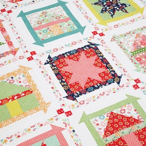 Pode incluir: Uma colcha de retalhos colorida com um padrão repetitivo de quadrados. Cada quadrado apresenta um design de estrela ou casa em tecidos florais vermelhos, rosa, verdes e amarelos. A colcha tem um fundo branco com detalhes florais.