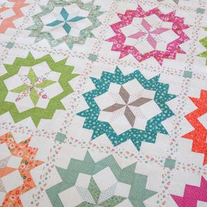 Könnte beinhalten: Eine Patchwork-Decke mit einem sich wiederholenden Sternmuster in verschiedenen Farben wie Pink, Grün, Orange und Türkis. Die Decke besteht aus weißen Stoffblöcken mit Blumen- und Vichy-Karomustern und zeigt aufwendige Steppnähte.