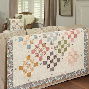 May include: A white quilt with a patchwork design featuring various floral patterns in shades of pink, blue, green, and brown. The quilt is displayed on a couch with a floral patterned border.