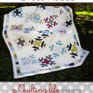 May include: A white quilt with a geometric pattern in shades of black, gray, red, teal, and green. The quilt is folded and lying on a bed of green grass. The quilt is titled "Cross Roads" and is a quilting pattern by "a Quilting life designs" by Sherri McConnell.