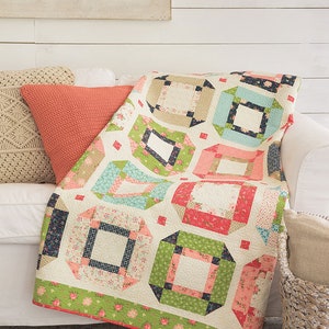 May include: A patchwork quilt with a geometric design in shades of pink, green, blue, and white. The quilt is made with a variety of fabrics, including floral prints and solid colours. It is displayed on a white sofa with a throw pillow.