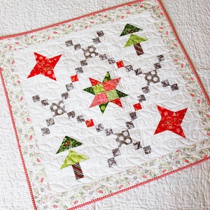 May include: A white Christmas-themed quilt with red and green accents. The quilt features red stars, green Christmas trees, and a central star design. The border has a floral pattern with a red and white striped edge. The quilt is approximately 30 inches square.