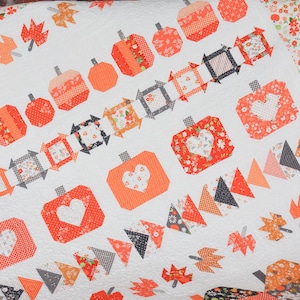 May include: A white quilt with a repeating pattern of orange pumpkins, hearts, and fall leaves. The quilt features various fabrics in orange, gray, and floral patterns. The quilt is a cozy, handmade item.