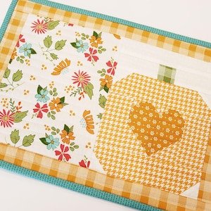 May include: A quilted placemat featuring a floral pattern with orange, blue, and pink flowers on a white background. A pumpkin-shaped design with a heart is on the right, set against a yellow and white houndstooth pattern. The border is yellow gingham with a teal edge.