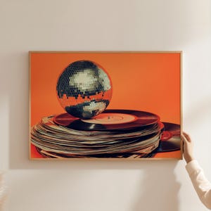 Puede incluir: Una bola de discoteca plateada se encuentra encima de una pila de discos de vinilo sobre un fondo naranja.
