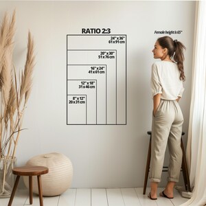 Op de afbeelding: Een witte muur met een zwart-witte grafiek die verschillende formaten van kunstprints in inches en centimeters laat zien. De grafiek is getiteld "Ratio 2:3". Een vrouw staat naast de grafiek, en de tekst "Female height is 65" staat boven haar.