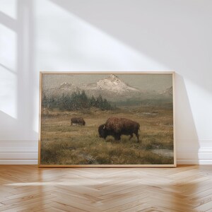 May include: A framed landscape painting depicts two bison grazing in a grassy field, with a mountain range in the background. The artwork features a muted color palette of browns, greens, and grays, creating a vintage aesthetic. The painting is displayed in a light-colored wooden frame.
