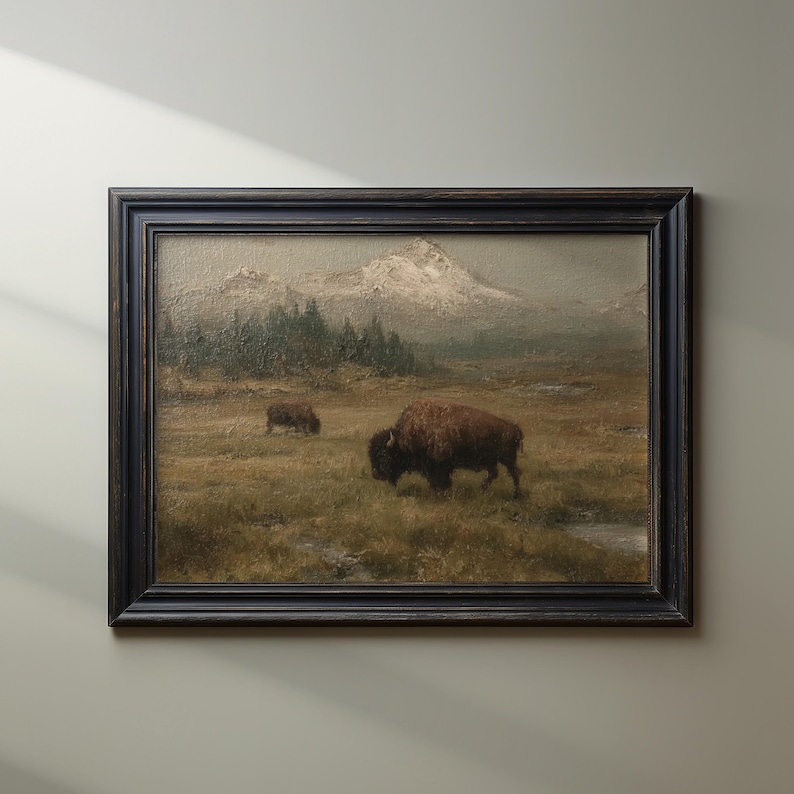 May include: An oil painting in a dark wood frame depicts two bison grazing in a grassy field. Mountains with snow-capped peaks are in the background. The artwork has a vintage aesthetic.