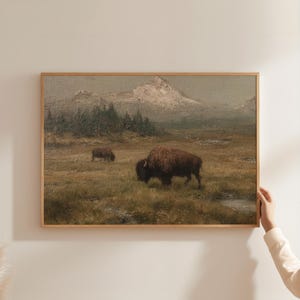 May include: A framed landscape painting depicts two bison grazing in a grassy field, with a mountain range in the background. The artwork features muted tones of brown, green, and gray, creating a serene, natural scene.