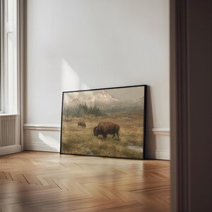 May include: Framed landscape art featuring two bison in a grassy field with mountains in the background. The artwork has a vintage aesthetic with muted colors and a black frame. The bison are brown and the mountains are snow-capped.