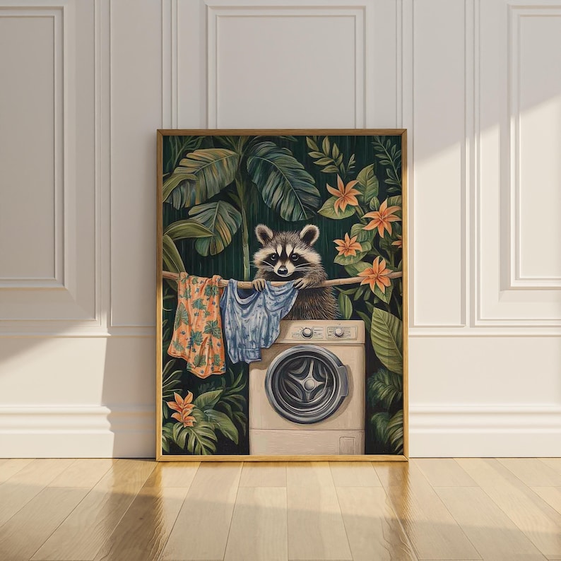 May include: A framed art print depicts a raccoon doing laundry. The raccoon is perched above a washing machine, surrounded by tropical foliage and orange flowers. Clothes hang on a clothesline. The artwork has a gold frame.
