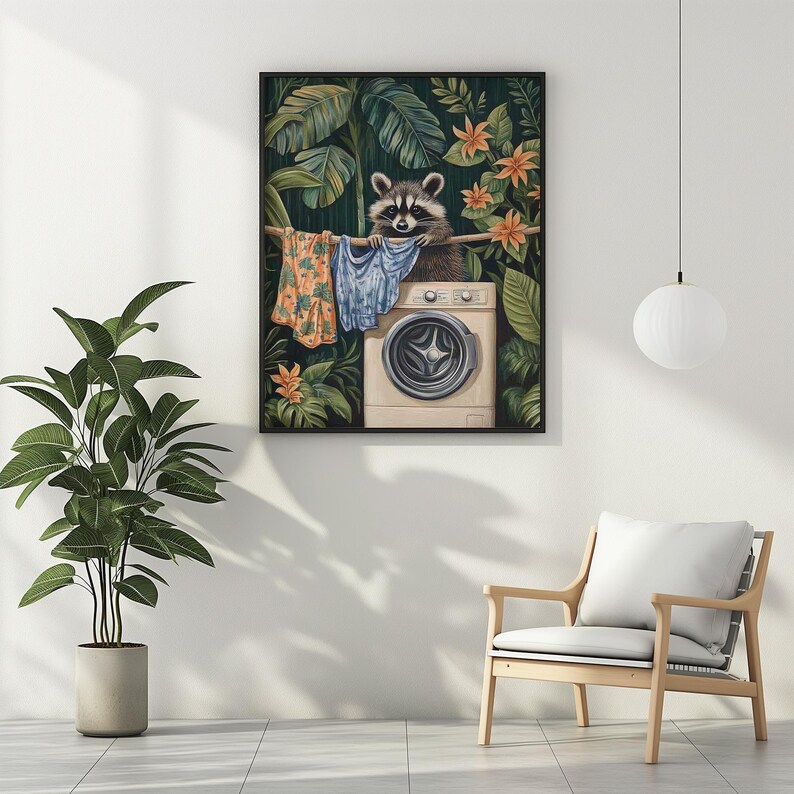 May include: A framed art print depicts a raccoon hanging laundry on a clothesline above a washing machine. The raccoon is surrounded by lush green foliage and orange flowers. The clothesline holds colorful shirts.