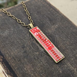 May include: A rectangular pendant necklace featuring a red circuit board design with white details and text. The pendant is encased in a gold-colored frame and hangs from a matching chain. The necklace is displayed on a dark, textured surface.