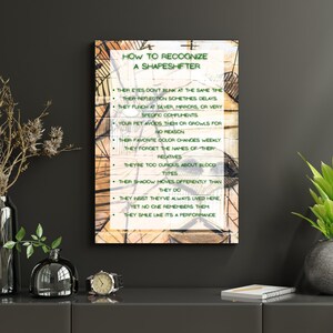May include: A framed artwork with the text "HOW TO RECOGNIZE A SHAPESHIFTER" and a list of characteristics. The artwork has a distressed, vintage appearance with a light brown background and green text. The frame is a light brown color.