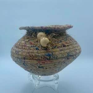 May include: A woven basket with a beige, brown, and blue pattern. The basket has a round shape and is decorated with white beads and brown string. The basket is sitting on a clear glass stand.
