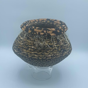 May include: A small, round, woven basket with a black and tan patterned fabric exterior. The basket has a decorative trim around the top and a metal heart charm hanging from the side.