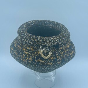May include: A black and gold fabric pot with a heart-shaped embellishment. The pot has a unique, rounded shape and a textured surface.