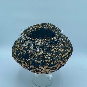 May include: A small, round, black and orange woven basket with a white and silver trim. The basket is sitting on a clear glass stand.