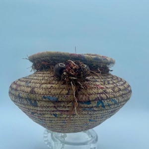 May include: A small, round, woven basket with a brown, beige, and blue pattern. The basket has a decorative trim around the top with brown beads and a brown tassel.