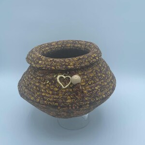 May include: A brown woven basket with a gold heart charm and a white bead hanging from the rim. The basket has a swirling pattern and is sitting on a clear stand.