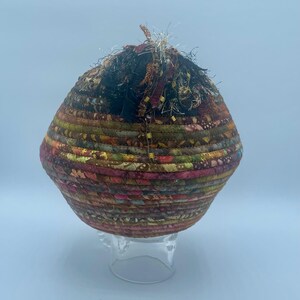 May include: A round, woven basket with a brown, green, and pink pattern. The basket is made of fabric scraps and has a frayed edge. It is sitting on a clear stand.