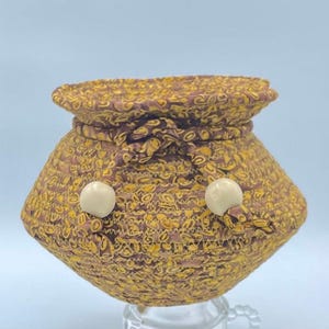 May include: A small, brown and gold patterned woven basket with white button accents. The basket has a round base and a slightly flared top.