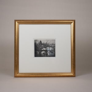 May include: A framed black and white etching of a cityscape, featuring a bridge and buildings. The artwork is set within a gold-colored frame and a white mat.