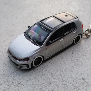 May include: A silver miniature car keyring with a black roof and black wheels. The small car has a detailed design, including windows and a red accent on the front. The keyring is attached to a silver ring.
