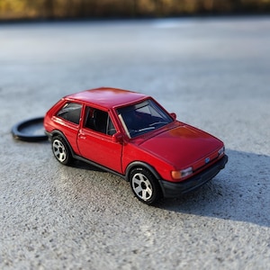 Puede incluir: Un pequeño coche de juguete rojo con ruedas negras y un llavero negro. El vehículo en miniatura tiene un diseño clásico de hatchback, con un acabado brillante. El coche está sobre un fondo borroso de árboles y cielo azul. Mide unos 10 cm de largo.