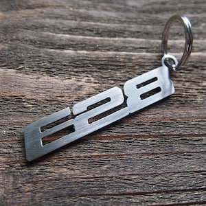 May include: A silver metal keyring featuring the number "828" in a cut-out design. The keyring is attached to a silver ring and lies on a rustic, weathered wooden surface. The design is simple and modern.