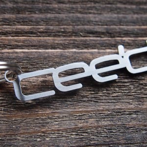 Peut inclure: Un porte-clés en métal argenté avec le mot "CEED" découpé dans une police de caractères moderne. Le porte-clés est doté d'un anneau en métal argenté.