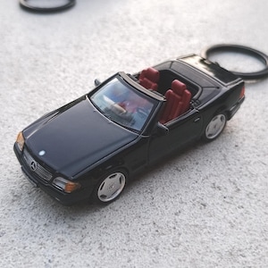 Puede incluir: Un llavero de coche descapotable negro con interior rojo. El coche en miniatura tiene ruedas plateadas y un logotipo de Mercedes-Benz en la parte delantera. El anillo del llavero es negro.