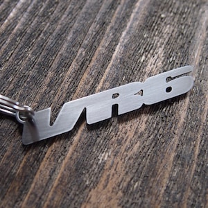 Puede incluir: Un llavero de metal plateado con las letras "VR6" recortadas. El llavero está unido a un pequeño anillo de metal. El fondo es una superficie de madera oscura y texturizada.