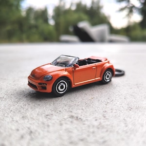 Könnte beinhalten: Ein leuchtend orangefarbenes Cabriolet-Spielzeugauto aus Druckguss mit schwarzem Innenraum und silbernen Felgen. Das Fahrzeug verfügt über eine klare Windschutzscheibe und einen kleinen Metallring an der Rückseite, der als Schlüsselanhänger dient.