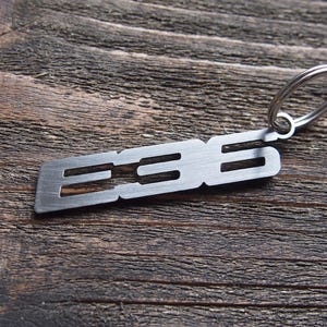 May include: A silver-colored metal keychain with the letters "E36" cut out. The keychain is rectangular with rounded edges and attached to a silver split ring. The background is a dark, textured wooden surface.