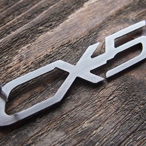 Puede incluir: Un llavero de metal plateado con las letras "CX5" recortadas. El llavero tiene un anillo.