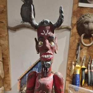 May include: A red wooden articulated doll with horns, a painted face, and white markings. The doll is wearing a brown fabric loincloth and is standing on a wooden base.