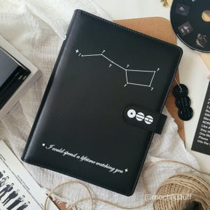 May include: Black leather journal with a white constellation design and the text "I could spend a lifetime watching you." The journal has a snap closure and the letters "ooo" on the strap.