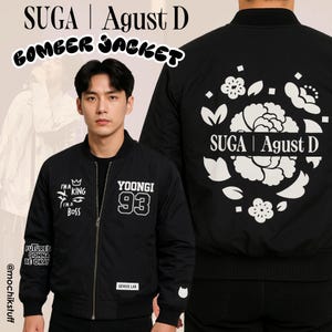 May include: Black bomber jacket with white text and graphics. The front features "I'M A KING I'M A BOSS" and "YOONGI 93". The back displays "SUGA | Agust D" with floral designs. The jacket has a zipper closure and ribbed cuffs.