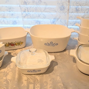 Może przedstawiać: Kolekcja białych naczyń CorningWare w stylu vintage z niebieskimi wzorami kwiatowymi. Zestaw zawiera różne rozmiary naczyń do zapiekania, niektóre ze szklanymi pokrywkami, idealne do pieczenia i serwowania. Naczynia są ułożone na wzorzystej powierzchni.