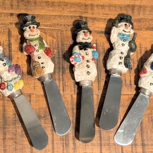 Può includere: Cinque spalma burro decorativi a forma di pupazzo di neve con lame in acciaio inossidabile. Ogni pupazzo di neve ha un design unico, indossa cappelli, sciarpe e tiene in mano vari oggetti a tema invernale. I manici sono dipinti con colori vivaci.