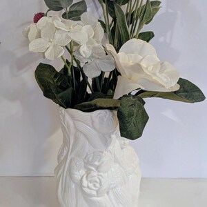 May include: A white ceramic vase with a floral design, filled with an arrangement of white and burgundy flowers and green leaves. The vase has a textured surface with embossed roses and foliage.