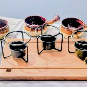 Op de afbeelding: Een set van zes keramische soepkommen met handvatten, in bruin- en crèmetinten, gerangschikt op een houten serveerplank. Elke kom wordt gecombineerd met een zwarte metalen standaard en een kleine zwarte metalen pot. De kommen hebben een donkerpaars interieur.