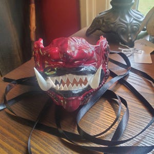 May include: A red and black Oni mask with white fangs and a wide, toothy grin. The mask has a textured surface and is tied with a black ribbon. The mask is a traditional Japanese demon mask.
