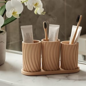 May include: A bathroom scene featuring a potted orchid with white flowers and a set of three beige toothbrush holders. Each holder contains a toothbrush and a tube of toothpaste. The holders sit on a matching tray.
