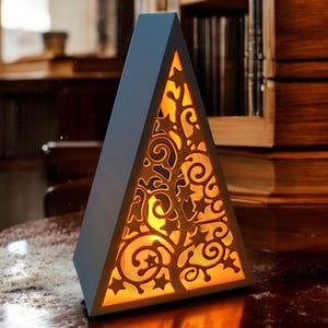 May include: A blue, pyramid-shaped lamp with a decorative, openwork design. The lamp's front panel features a cut-out pattern of swirling lines and star shapes, with a warm, yellow light emanating from within. The lamp is placed on a wooden surface.