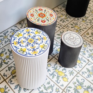 Op de afbeelding: Een verzameling decoratieve keramische krukjes met patronen op de bovenkant. Eén krukje is wit met een blauw en geel citroenontwerp, twee zijn donkergrijs met bloempatronen en één is zwart. De krukjes hebben een geribbelde textuur.