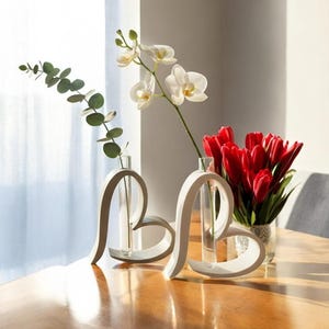 Op de afbeelding: Twee witte hartvormige frames, elk met een doorzichtige glazen reageerbuisvaas. Eén vaas bevat een groene eucalyptusstengel, terwijl de andere een witte orchideetak bevat. Een aparte glazen vaas bevat een boeket met rode tulpen.